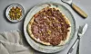 Dessertpizza med sjokoladepålegg og pistasjnøtter på en tallerken sett ovenfra. Sprø dessertpizza toppet med sjokoladepålegg, hakkede pistasjnøtter og flaksalt servert på fat med nøtter ved siden av.
