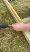 Hånd som bruker drill til å skru i en trelekt på gressplen med tommestokk ved siden av.