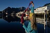 Person ved kai løfter en rosa strikket veske, iført gult hekleskaut og blå genser foran stille fjord.
