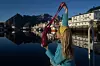 Person ved kai løfter en rosa strikket veske, iført gult hekleskaut og blå genser foran stille fjord.