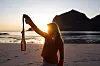 Person på strand i solnedgang holder et heklet flaskenett med flaske.