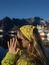 Person ved vannkant med gult heklet skaut og grønn genser foran fjell og hus.