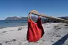 Rødrosa håndlaget veske med bambushåndtak henger på en pinne ved en sandstrand.