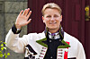Ung mann i bunad smiler og hilser med hånden hevet foran en bygning.