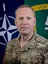Militæroffiser i kamuflasjeuniform foran Nato-flagg og grønt militært banner.