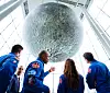 Artemis II-astronauter og Mark Carney ser opp på en stor månemodell i glasshall.