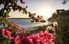 Solnedgang over rolig middelhavsstrand med rosa blomster i forgrunnen og få mennesker på promenaden.