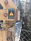 Nærbilde av advarselsskilt på trekonstruksjon ved en smal hengebru i skogsterreng.