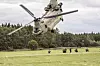 Et stort militærhelikopter flyr lavt over en grasmark med soldater på bakken og skog i bakgrunnen.