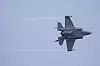 En grå F-35-jager flyr mot blå himmel med kondensstriper bak.