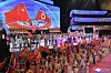 Stor gruppe mennesker med røde og oransje flagg på en arena med store skjermer og lysende scene.