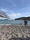 Strand med blåstripet parasoll, badende personer i vannet og kystby under klar himmel.