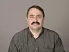 Mann med moustache sitter foran en grå bakgrunn.