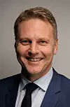 Smilende mann i blå dress og mørkt slips foran grå bakgrunn.