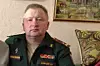 En mann i militæruniform sitter innendørs foran et mønstret veggteppe og en innrammet bilde.