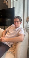 Ung mann i hvit polo sitter i en lys sofa ved et vindu innendørs.