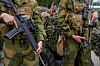 To soldater i kamuflasjeuniform holder hver sin rifle, med kjøretøy og camouflage-nett i bakgrunnen.