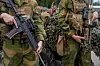 To soldater i kamuflasjeuniform holder hver sin rifle, med kjøretøy og camouflage-nett i bakgrunnen.