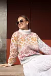 En kvinne sitter utendørs i en mønstret genser i hvitt, rosa og oransje med solbriller.
