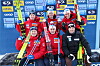 Seks langrennsløpere står og sitter foran en sponsorvegg med ski og vinterklær etter rennet.