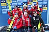 Seks langrennsløpere står og sitter foran en sponsorvegg med ski og vinterklær etter rennet.