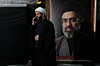 En mann i svart kappe og hvit turban snakker i mobiltelefon ved siden av et stort portrett av Ayatollah Mojtaba Khamenei.