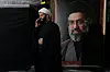 En mann i svart kappe og hvit turban snakker i mobiltelefon ved siden av et stort portrett av Ayatollah Mojtaba Khamenei.