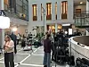 Filmteam og kamerautstyr i et lyst atrium med balkonger og høye vinduer.