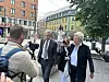 Personer samles på et bytorv mens fotografer og en kameramann filmer dem foran bygårder.