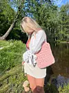 En kvinne i hvit kjole står ved en elv med en rosa heklet veske over skulderen.