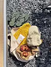 En heklet veske med strandting på grå rullestein, ved siden av grønne sandaler.