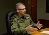 En mann i militæruniform sitter ved et bord og snakker mens han gestikulerer med hendene.