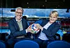 To personer sitter i blå stoler og holder en fotball sammen innendørs.