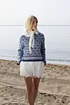 Kvinne i blåhvit mønsterstrikket genser og hvit shorts står på en sandstrand ved sjøen.