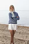 En kvinne står på en strand og viser frem en blå og hvit strikkegenser med mønster.