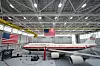 Et stort hvitt presidentfly står utstilt i en hangar med to amerikanske flagg.