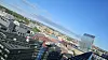 Skrått luftfoto av by med kontorbygg, takflater og en glasskledd høyblokk mot blå himmel.