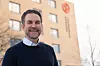Smilende mann foran et lysgult kontorbygg med logo og vinduer.