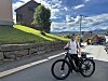 Person med svart sykkel i en bratt gate med hus, steinmur og solfylt himmel.