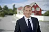 Mann i mørk dress står foran en vei med rødt hus og hvitt gjerde i bakgrunnen.