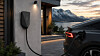 En svart elbil står parkert ved en veggmontert ladestasjon utenfor et hus ved solnedgang.