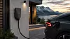 En svart elbil står parkert ved en veggmontert ladestasjon utenfor et hus ved solnedgang.