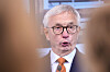 En mann med hvitt hår og briller taler foran en mikrofon, delvis rammet inn av uskarpe forgrunnsformer.