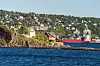 Kystlandskap med hus på en klippe, blått hav i forgrunnen og en rød lastebåt ved kaien.