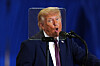 Donald Trump taler bak et beskyttelsesskjold foran mikrofoner mot blå bakgrunn.