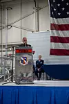 Speaker at a podium during a military retirement ceremony, with a seated uniformed attendee and a large U.S. flag behind.
