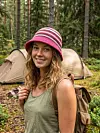Bildet viser en heklet Elton bøttehatt i rosa, bordeaux og ubleket hvitt, brukt på tur i skogen med telt i bakgrunnen. Bildet viser hvordan samme gratis hekleoppskrift kan få et roligere uttrykk med andre farger, og passer fint til hytte, camping og sommerdager ute.
