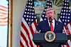 Donald Trump stands at a podium with the presidential seal, speaking indoors with U.S. flags behind him.