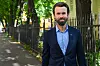 Mann i mørk dress står ved et gjerde langs en trebevokst gate.