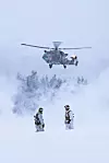 Helikopter henger lavt over snødekt terreng mens to soldater står i vinterlandskapet.